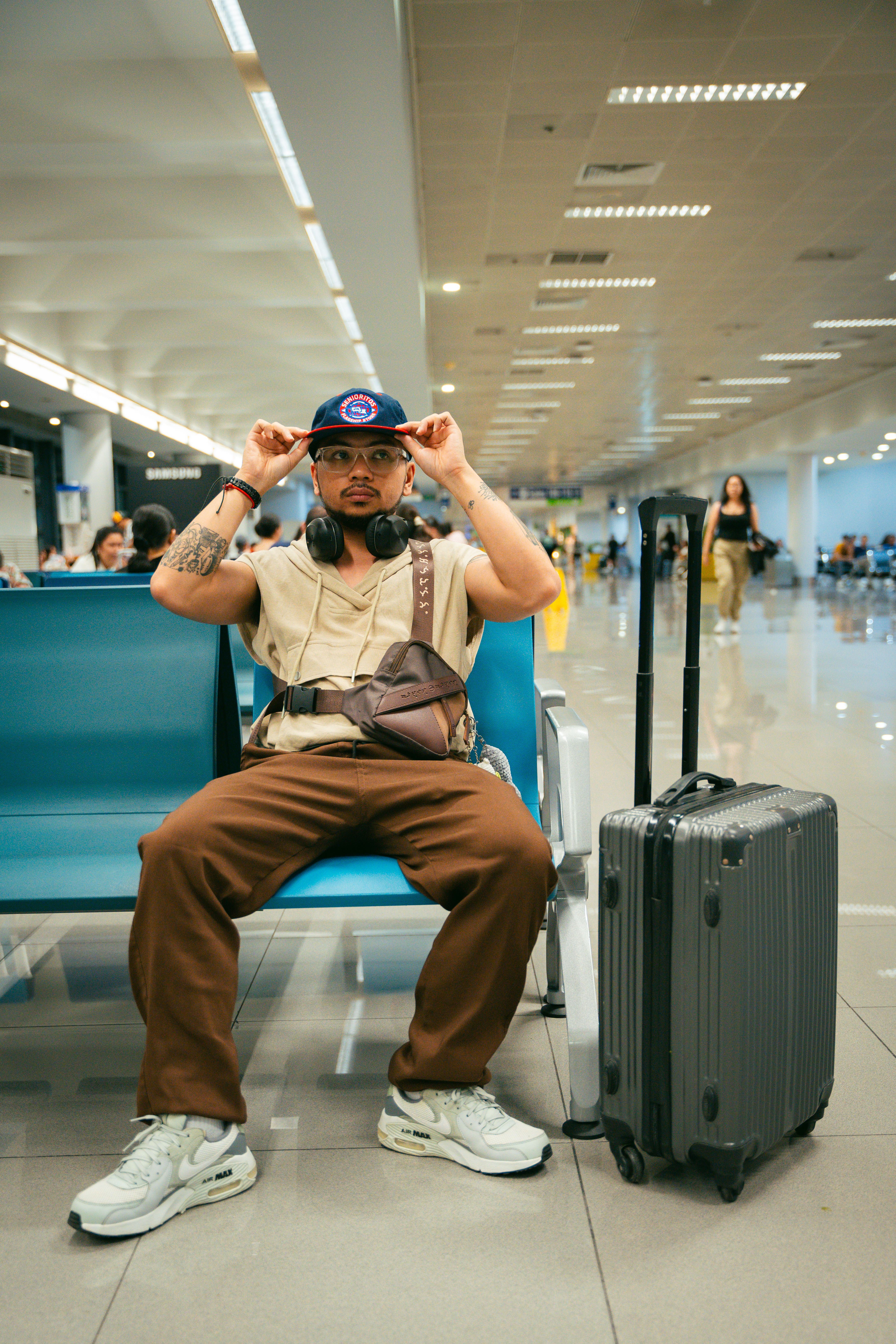A traveler waits in an airport beside a generic hard-shell suitcase; it is not identified as either carry-on compared here.