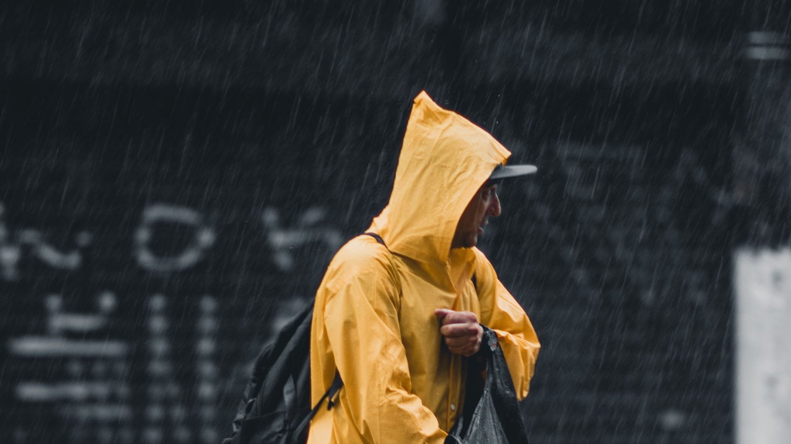 A commuter in a bright yellow raincoat walks through heavy rain on a city street; the photo illustrates outerwear and does not identify a product.