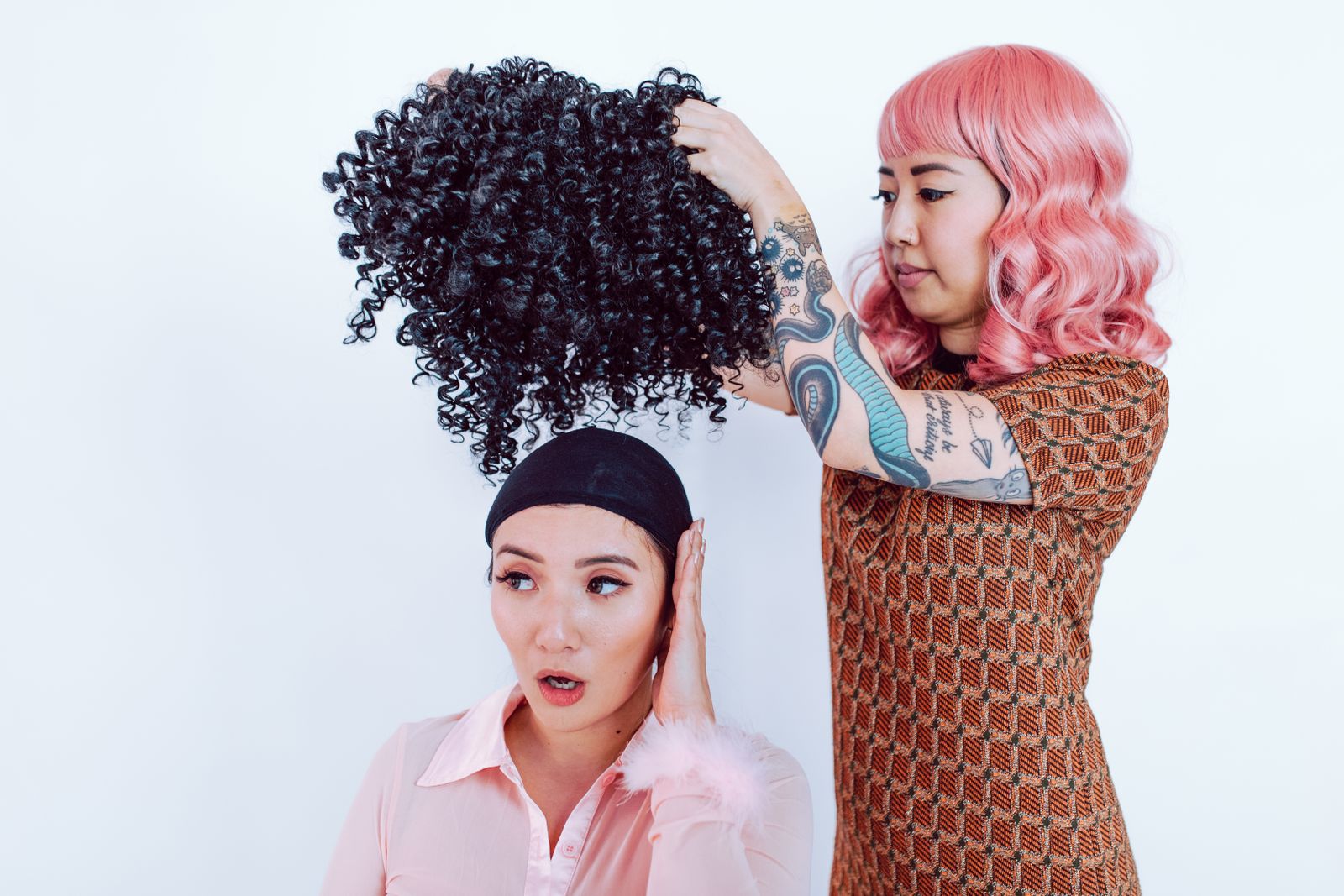 A stylist adjusts a curly wig on an adult client in a salon; the image is illustrative and does not identify the wig fiber.